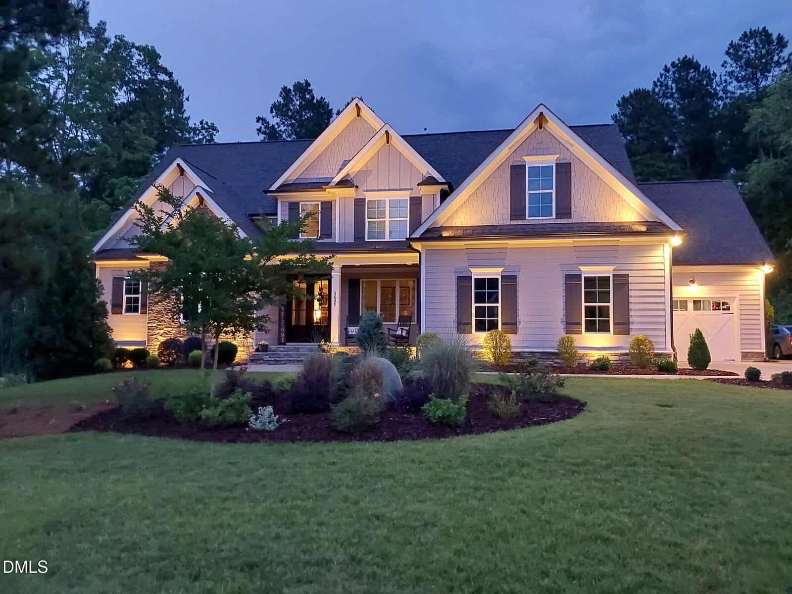 A large, modern two-story house with warm exterior lighting, multiple peaked roofs, and a well-maintained front yard with shrubs and trees, photographed at dusk.