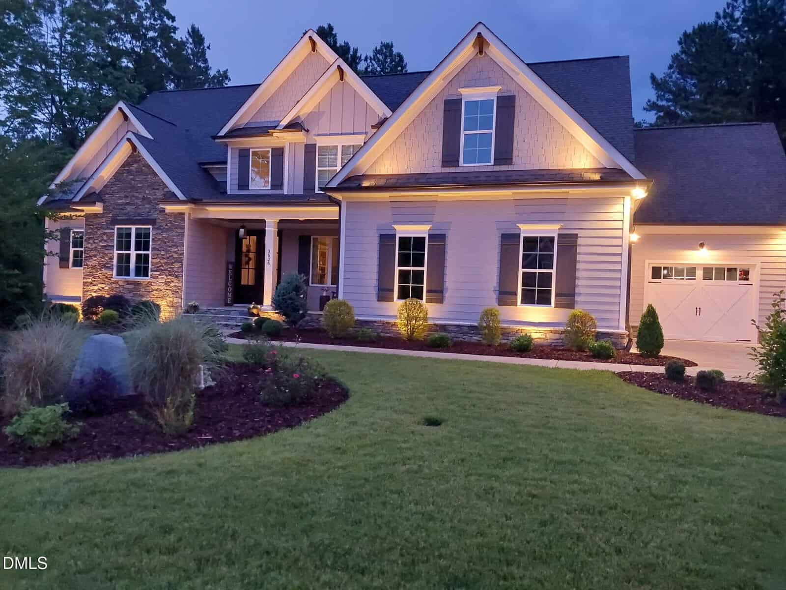 A modern suburban house with stone and siding exterior is warmly lit at dusk. The home has manicured landscaping, a covered front porch, and an attached garage, with trees and a lush green lawn in front.