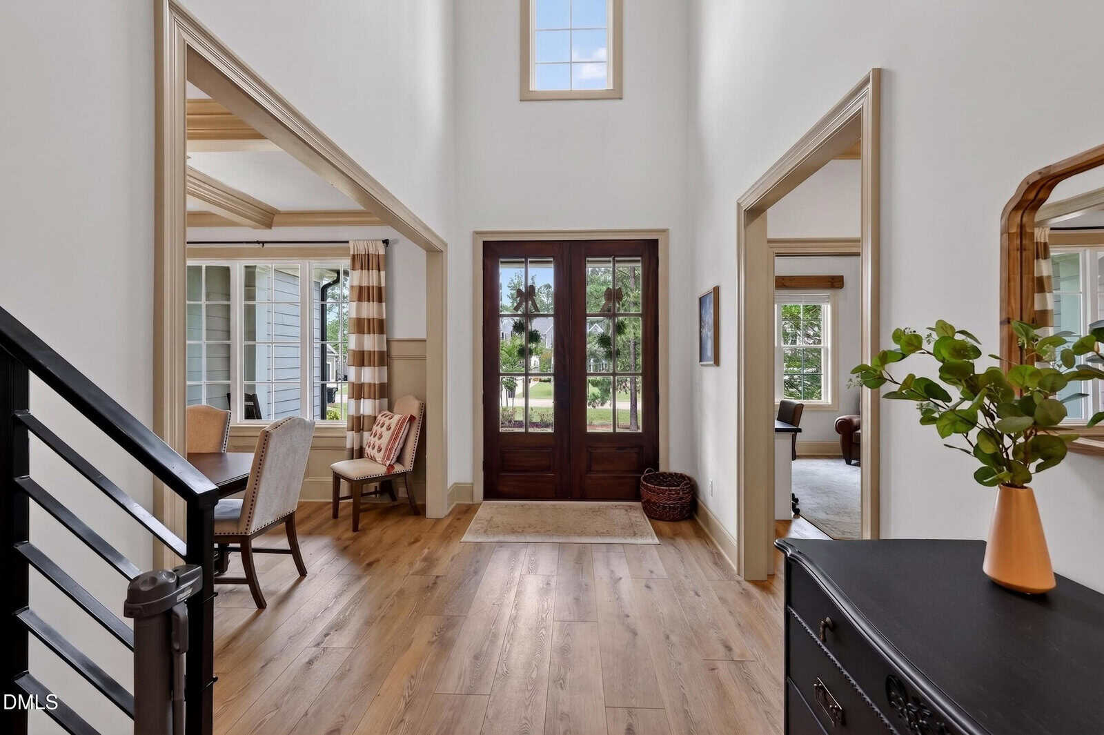 Bright entryway with wood floors, double front doors, a window above, and a view of rooms on both sides. A staircase with metal railing is on the left, and a black chest with a plant and mirror is on the right.