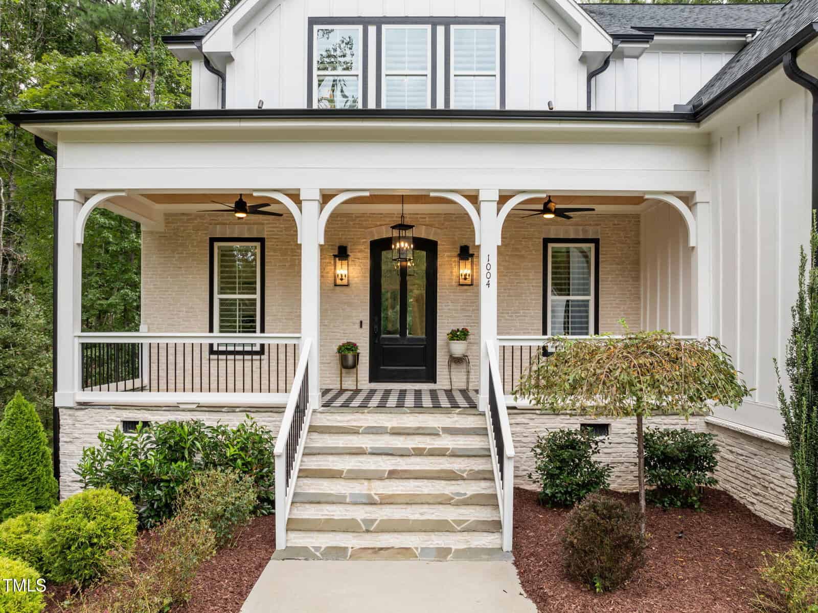 A modern farmhouse porch with a black front door, two hanging lantern lights, stone steps, white columns, black railings, and neatly landscaped plants along the walkway.