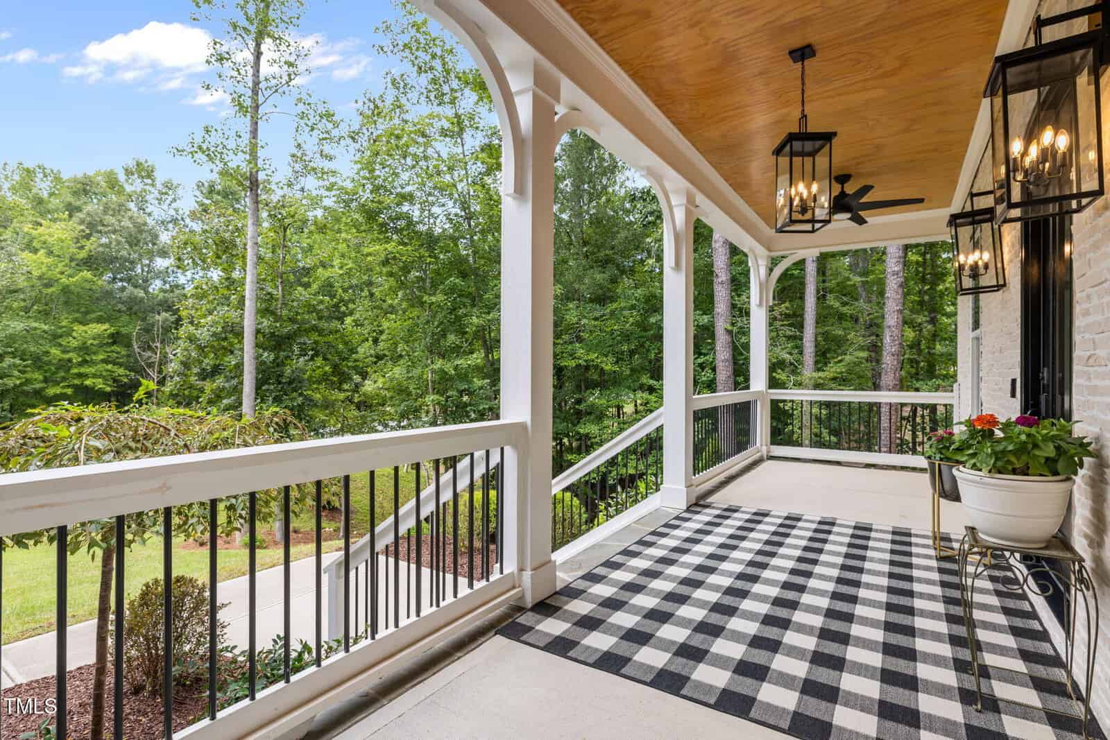 A covered porch with white railings and columns overlooks a green wooded area. The floor features a black-and-white checkered rug, potted flowers, and hanging lantern-style lights.
