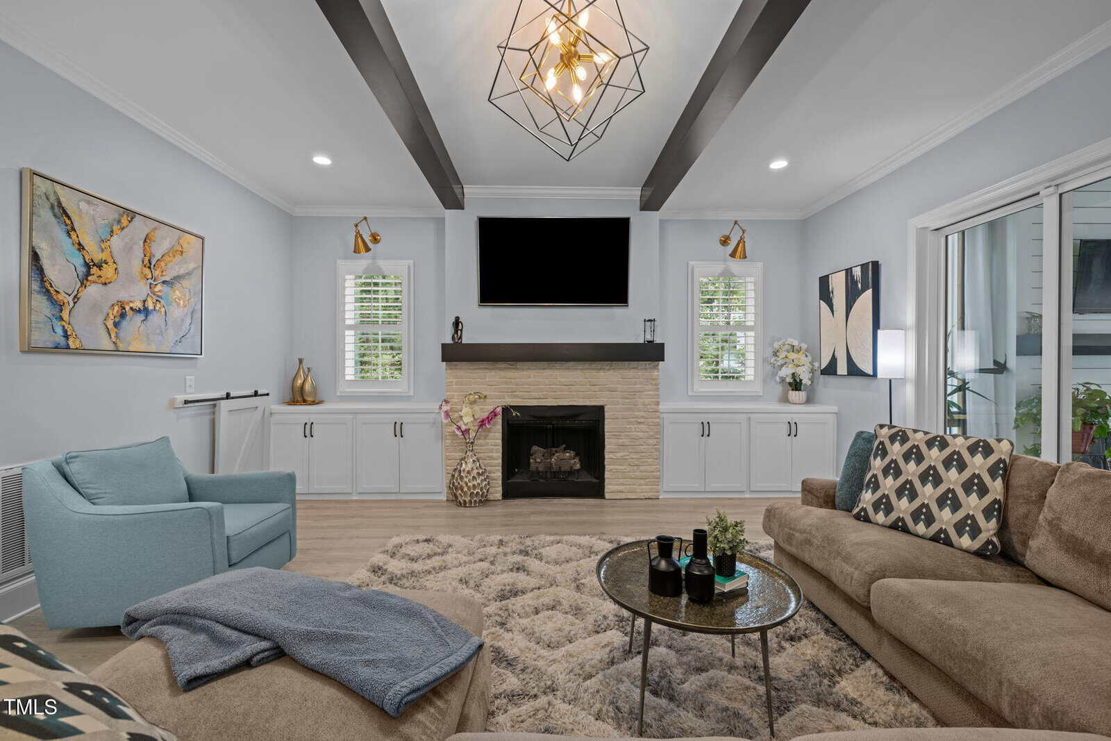 Modern living room with light blue walls, a beige couch, blue armchair, geometric chandelier, and a fireplace centered between two windows. A round coffee table sits on a patterned rug. Contemporary art decorates the space.