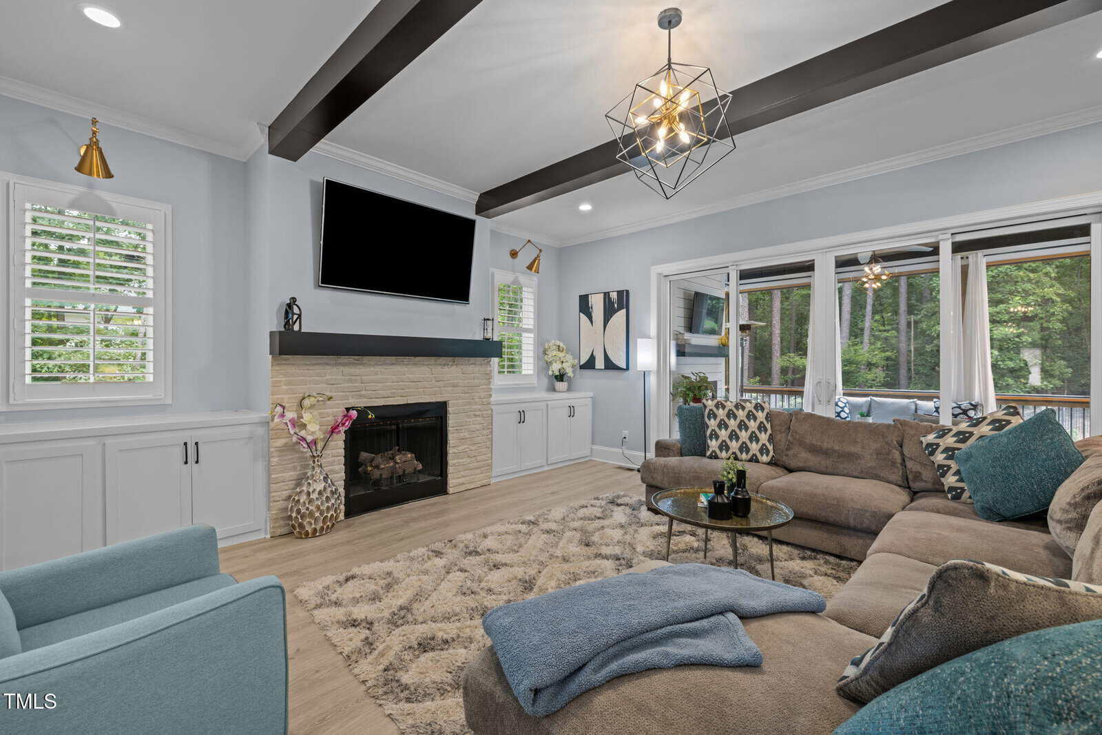 Modern living room with a sectional sofa, blue armchair, fireplace, large TV, rug, and glass coffee table. Large windows reveal a deck and forested area outside. Contemporary lighting and wood ceiling beams are featured.