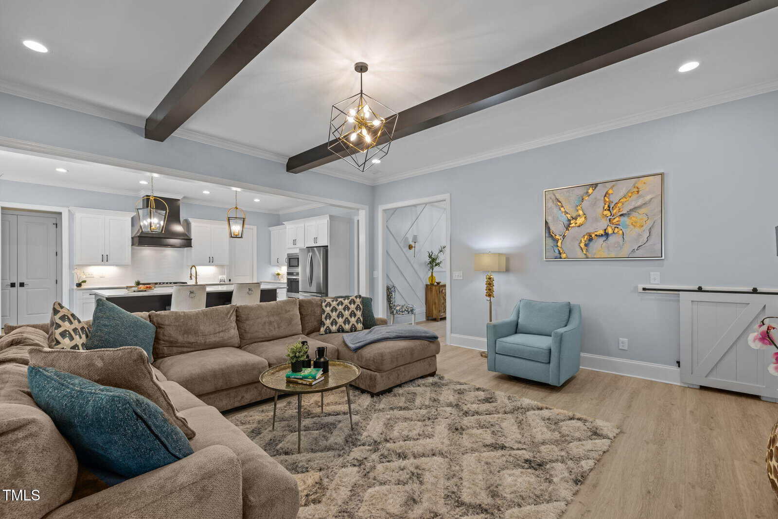 Modern living room with beige sectional, blue armchair, round coffee table, and patterned rug. Open kitchen in background, pendant lights, wood beams on ceiling, and abstract wall art above a lamp and small plant.