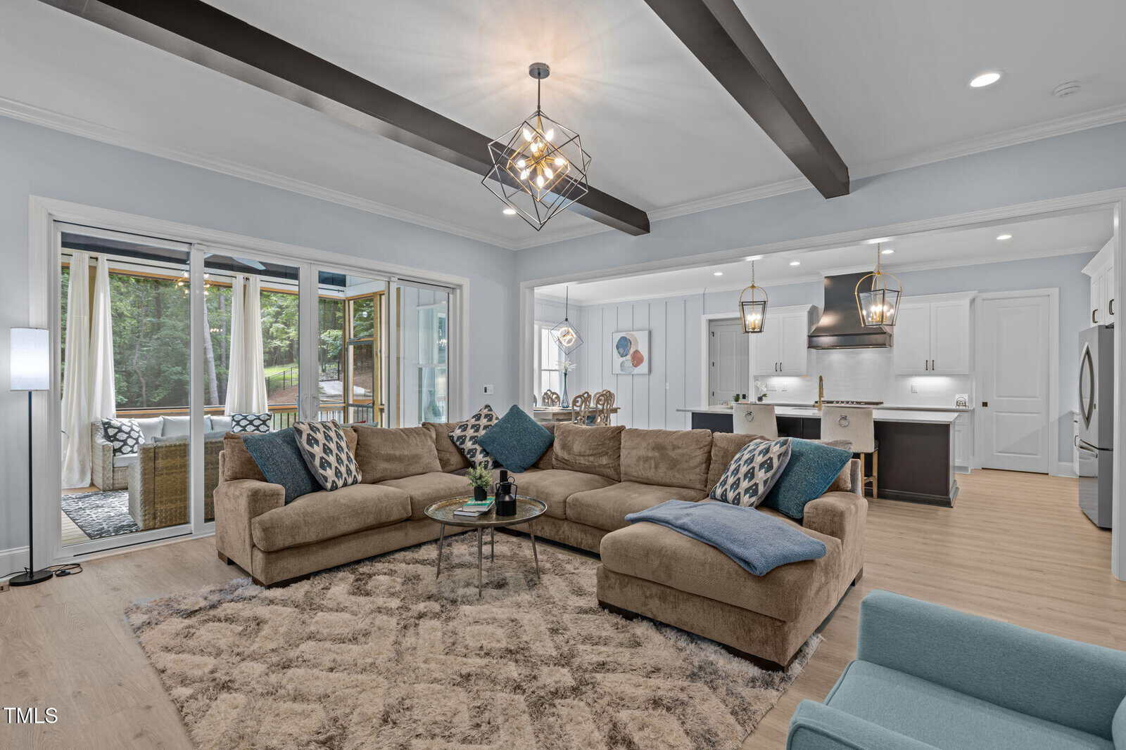 Modern open-concept living room with a brown sectional sofa, blue cushions, geometric chandelier, and a view of the kitchen with pendant lights and island seating. Sliding doors open to a sunroom with wicker furniture.
