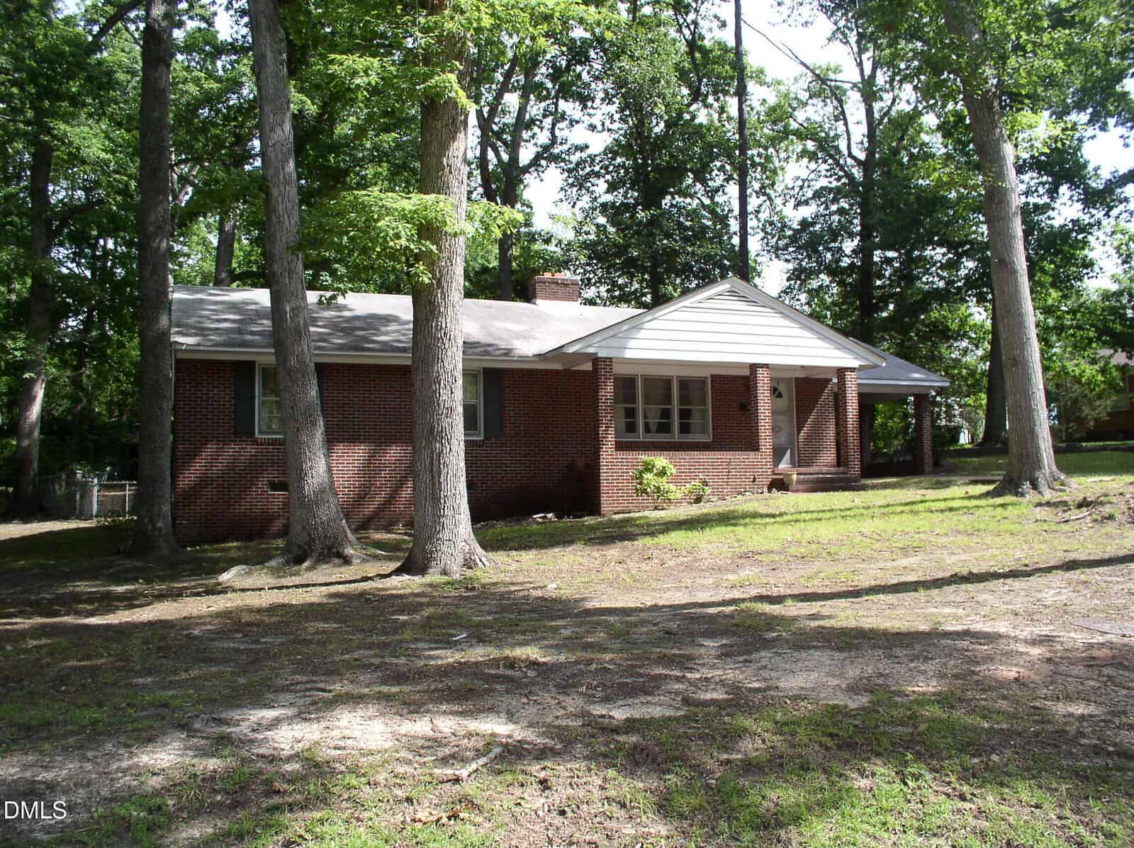 A single-story brick house with a gray roof sits among tall trees on a spacious, shaded yard at 1820 S Garnett Street Extension in Henderson. The home features a small covered front porch and several windows.