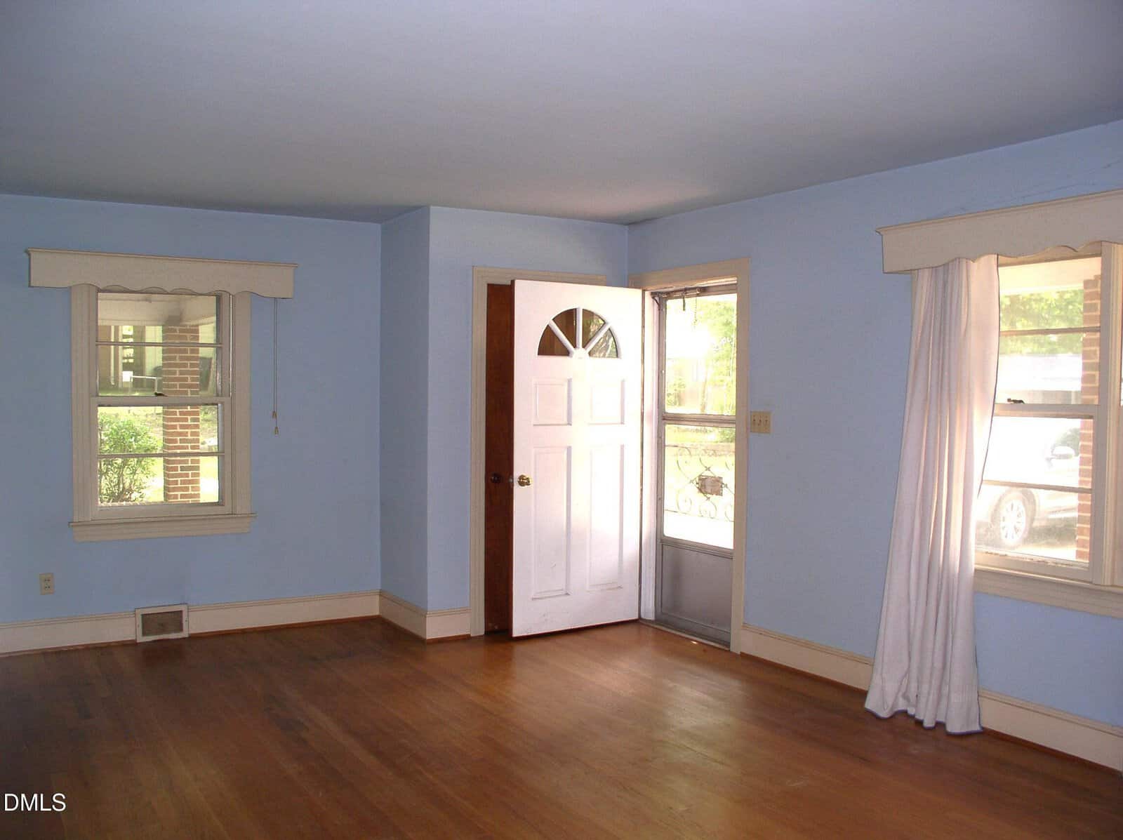 Empty living room with light blue walls, two windows with white valances, hardwood floor, a white front door, and a glass storm door. Sunlight streams in and white curtains hang by one window.