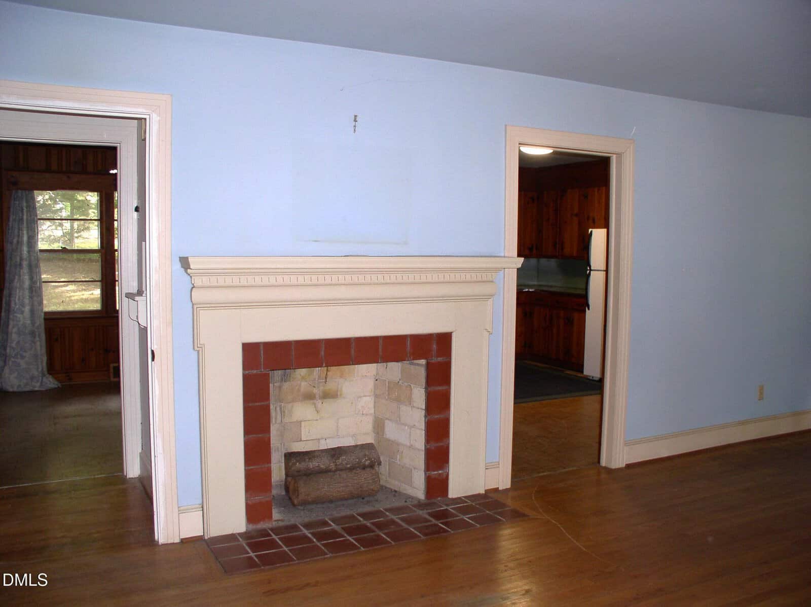 A room with light blue walls, a white fireplace with a brick opening, wood flooring, and two doorways leading to other rooms. One room has wood paneling; the other has a window with curtains.