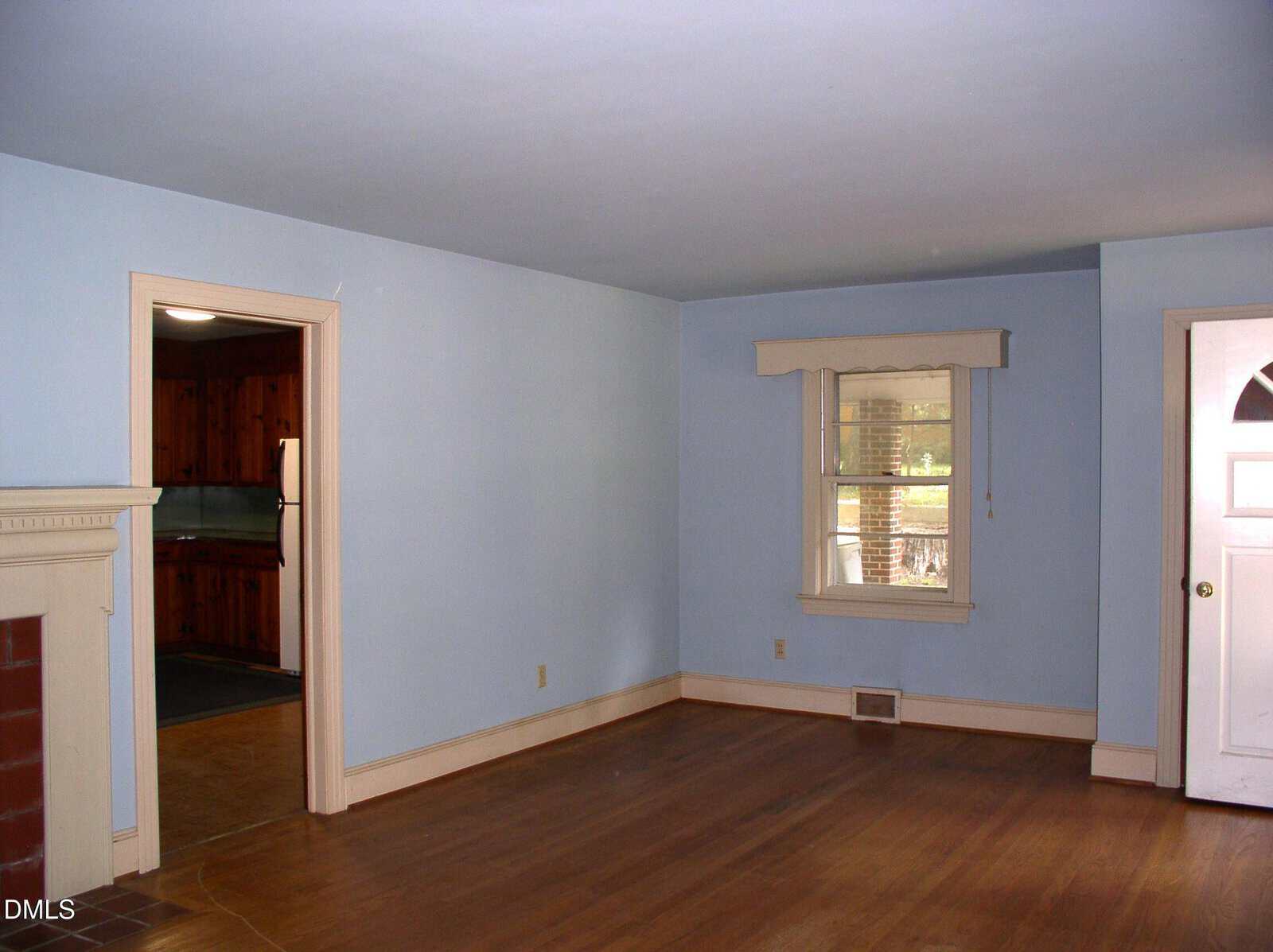 A mostly empty living room with light blue walls, white trim, hardwood floors, a window with a valance, a white front door, and an open doorway leading to a wood-paneled kitchen.