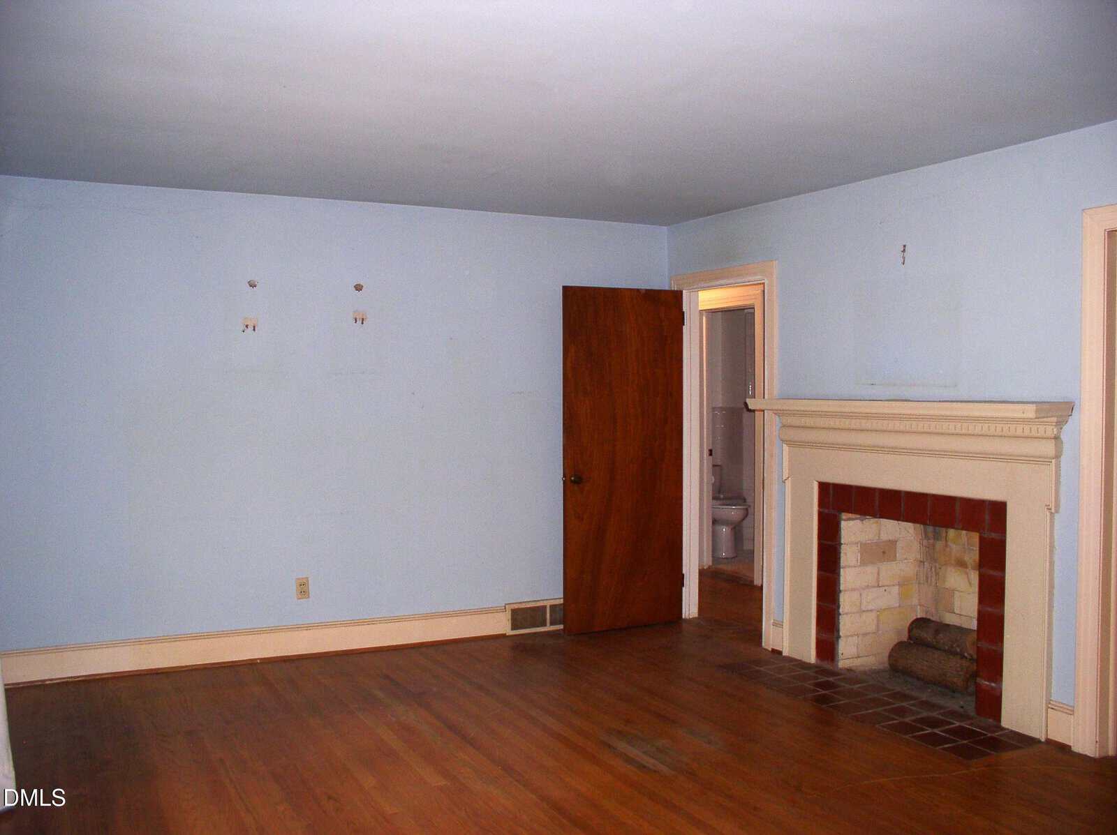 Empty room with light blue walls, hardwood floor, a wooden door, and a white fireplace with a red-tiled hearth. An open doorway reveals part of a bathroom in the background.