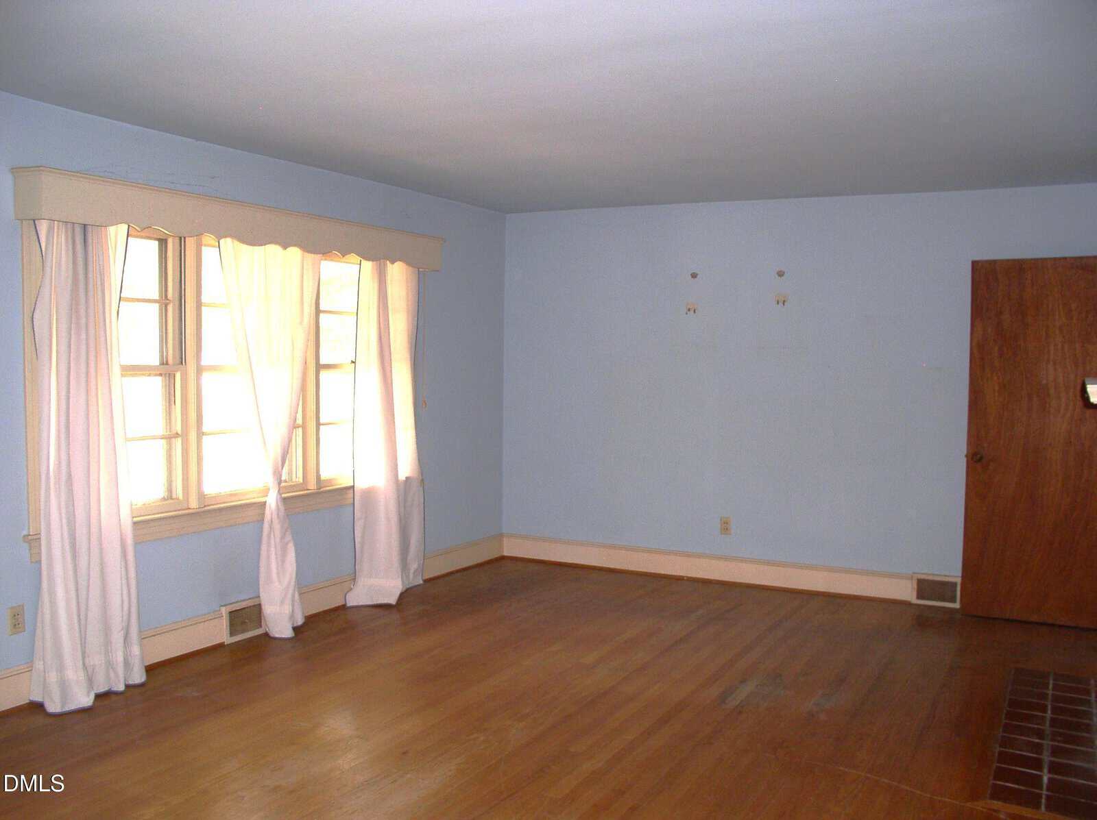 Empty room with bare hardwood floor, light blue walls, a large window with white curtains, baseboards, and a closed wooden door. The room is well-lit from natural light coming through the window.
