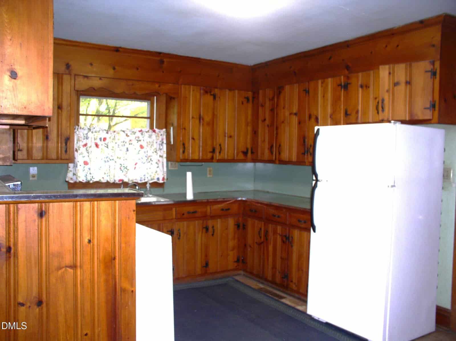 A kitchen with wood-paneled cabinets, a white refrigerator, a small window with floral curtains, light green walls, and a gray rug on the floor.