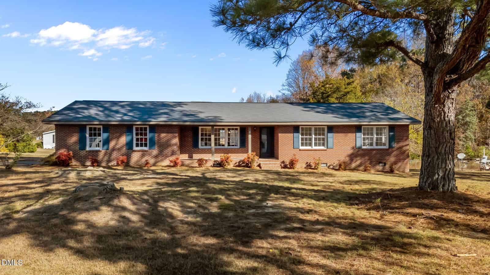 A single-story red brick house at 10 Alease Drive, Zebulon, features white-trimmed windows and a dark gray roof, set amid a grassy yard with small shrubs and a large tree in the foreground under a blue sky.