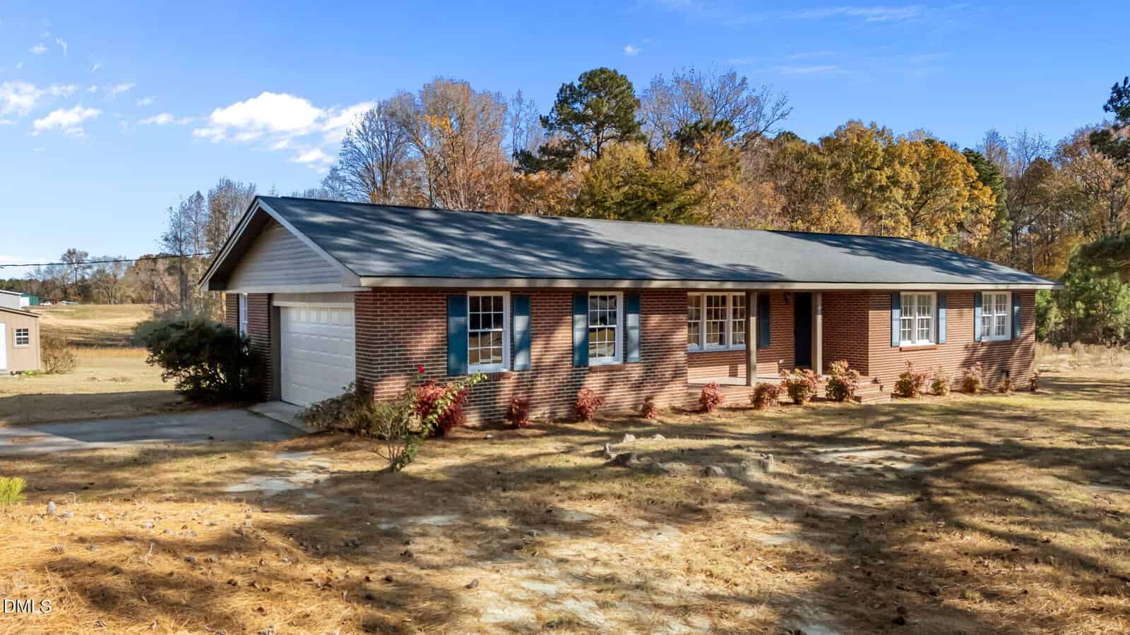 A single-story brick house with a gray roof, attached two-car garage, and blue-trimmed windows, surrounded by trees and dry grass under a clear blue sky.