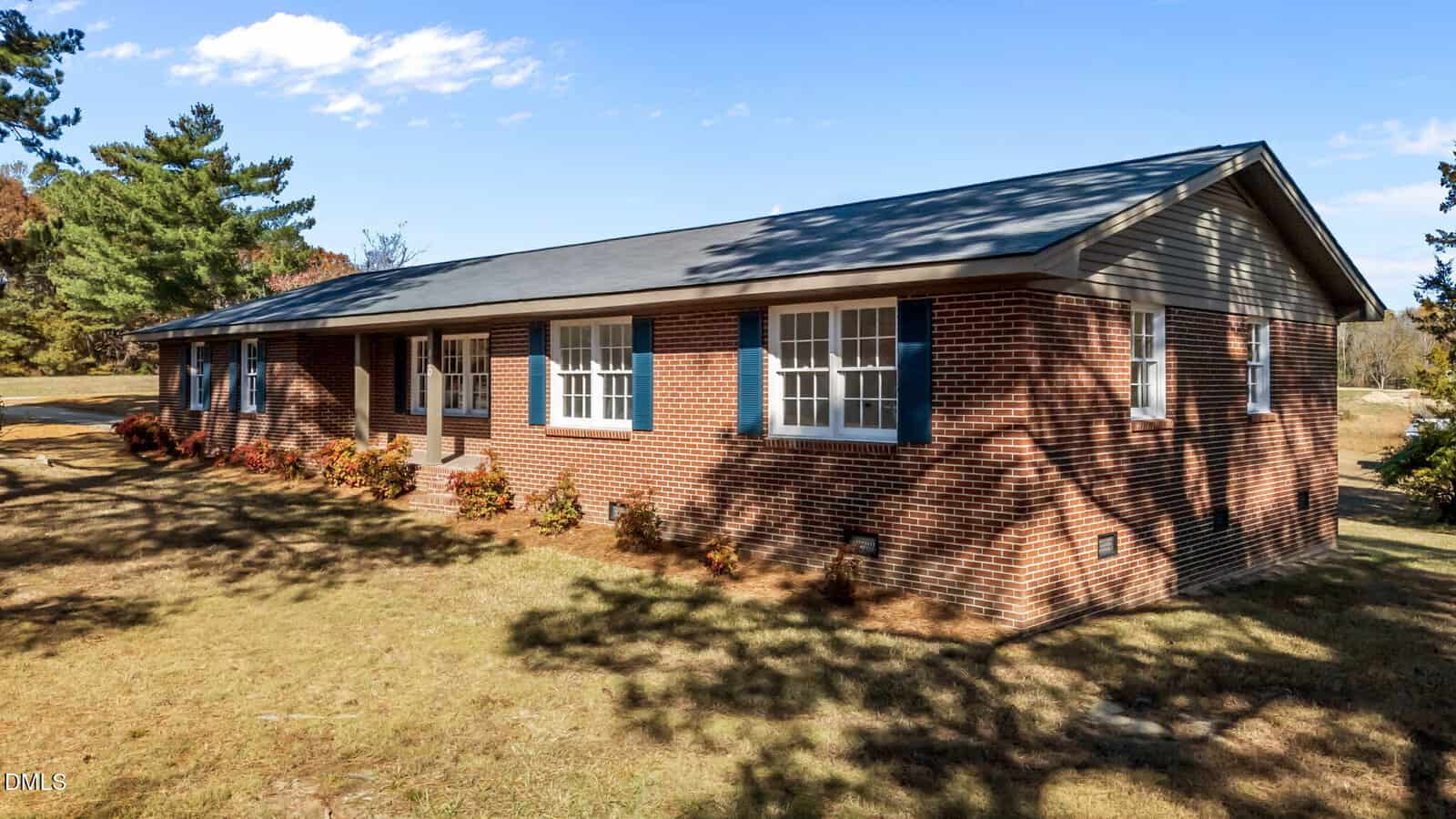 Single-story brick house with blue-gray shutters and a dark roof, surrounded by grass and trees, under a blue sky with scattered clouds. Shadows from nearby trees fall across the yard and the house.