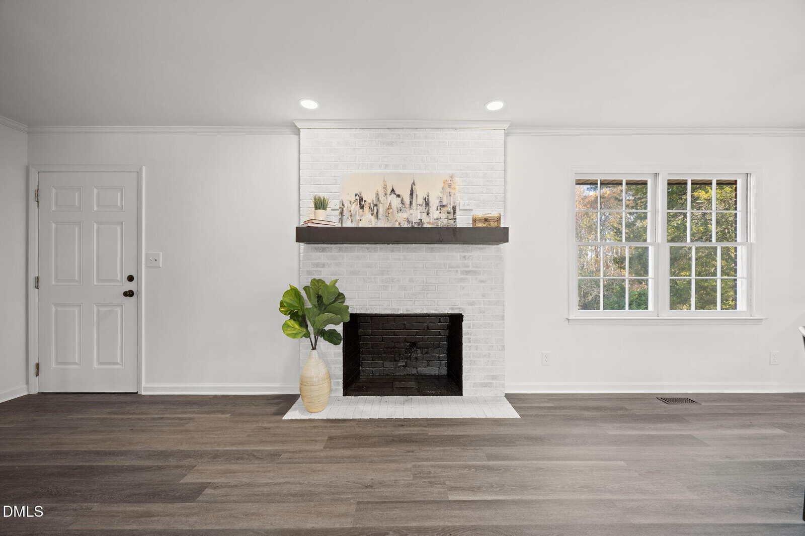 Modern living room with white walls and a white brick fireplace, dark wood mantel, large plant in a cream vase, and a window letting in natural light. The floor is dark wood, and there is a white door to the left.