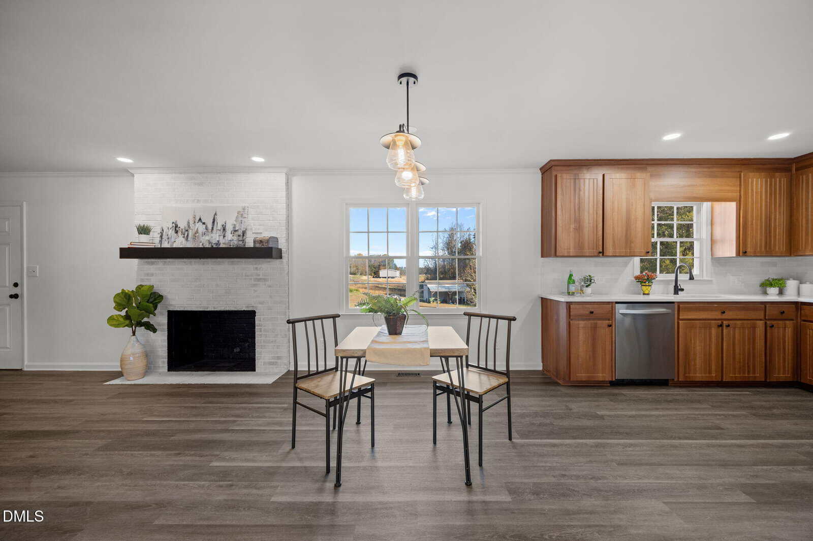 A dining table with four chairs sits in an open space between a white brick fireplace and a kitchen with wooden cabinets, stainless steel appliances, and a large window.