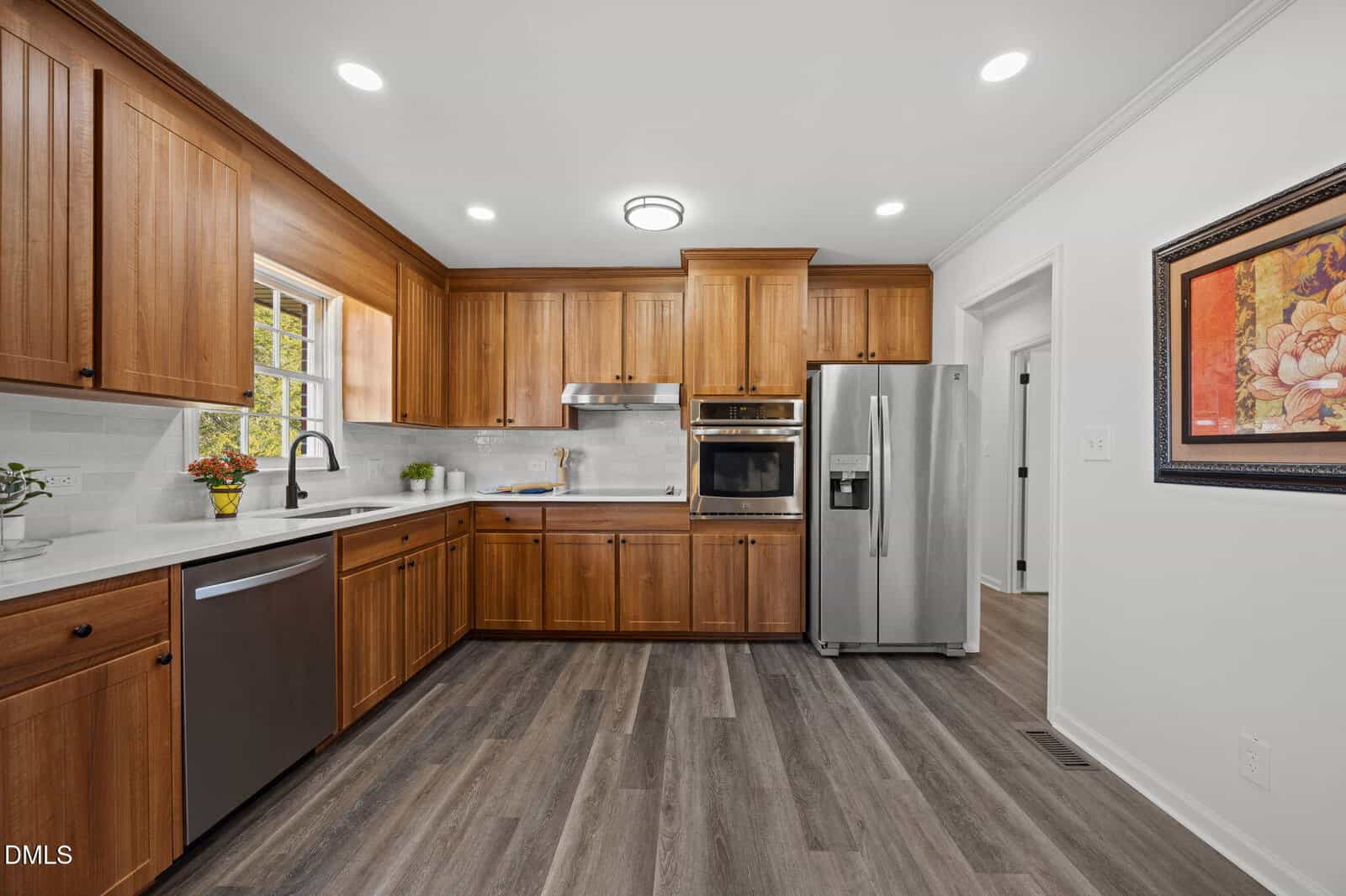 Modern kitchen with wooden cabinets, stainless steel appliances, white countertops, and a window above the sink. The floor is dark wood, and there is a framed floral artwork on the wall next to the refrigerator.