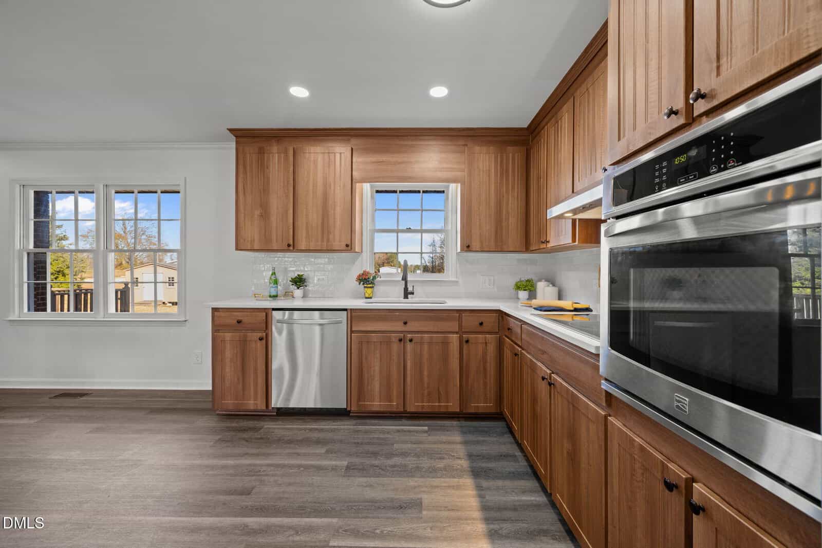 Modern kitchen with wooden cabinets, stainless steel appliances, double oven, dishwasher, sink under a window, white tile backsplash, and dark wood flooring. Sunlight enters through windows on the left and above the sink.