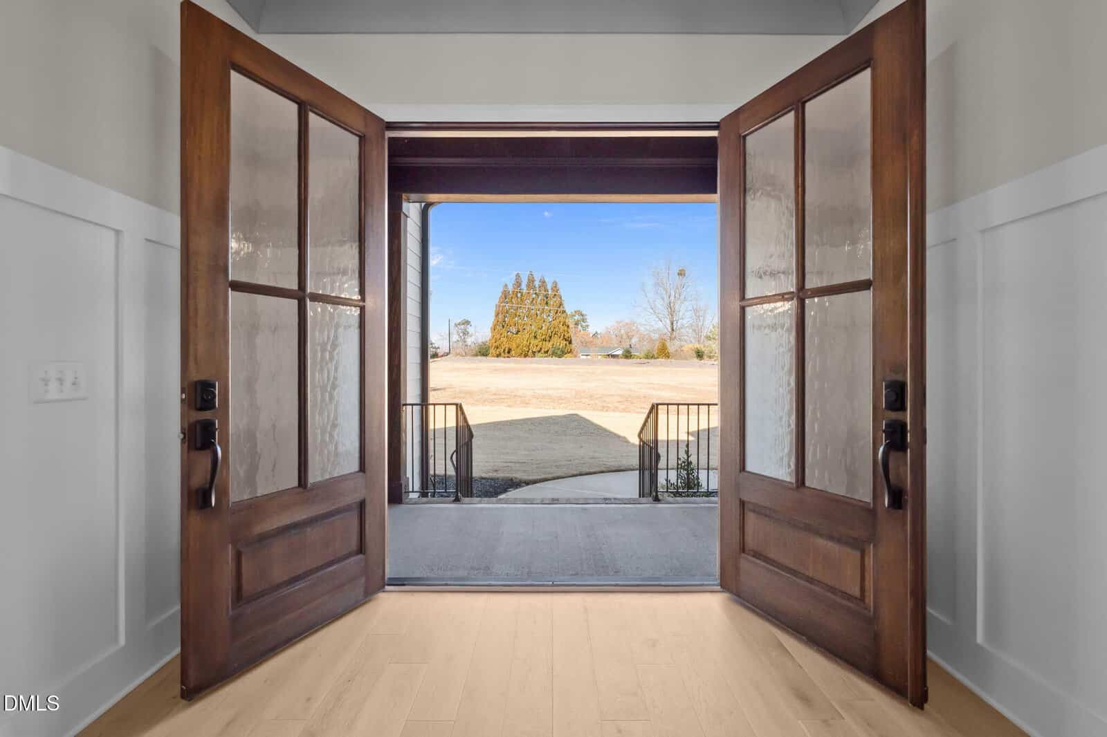Open double wooden doors reveal a porch and a view of a grassy yard with trees and a clear blue sky, seen from inside a bright entryway with light wood floors and white walls.