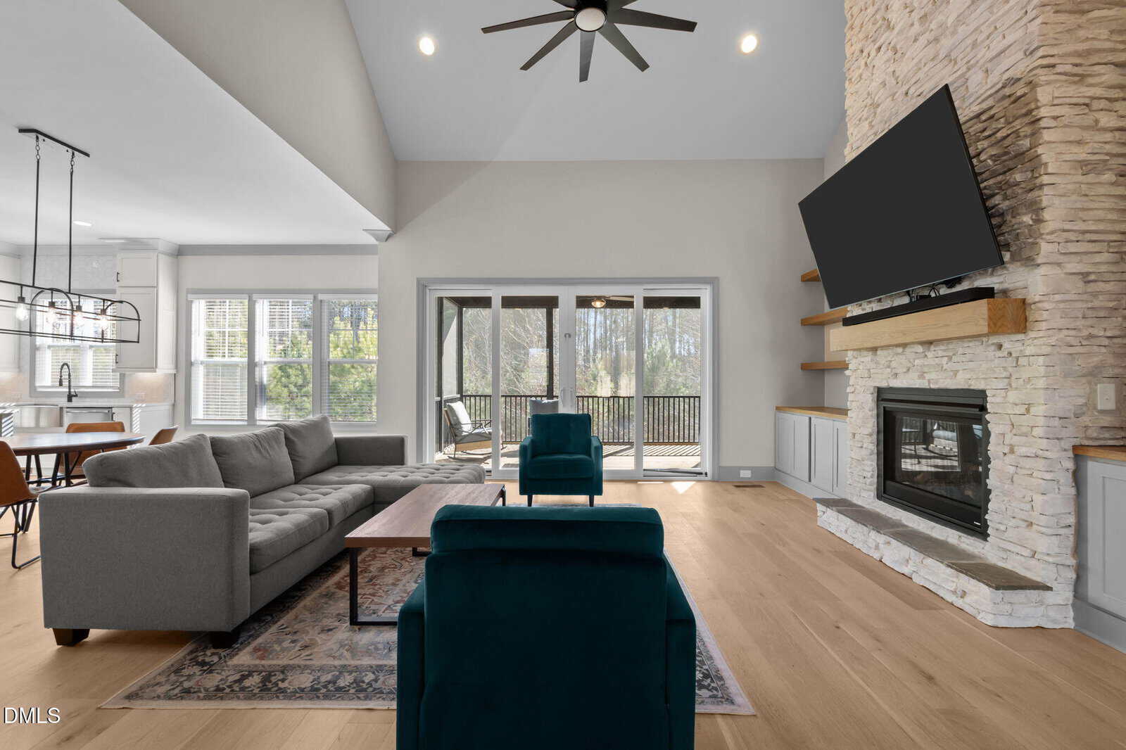 A modern living room with light wood floors, a stone fireplace, a mounted TV, gray sofa, two teal chairs, a wooden coffee table, and large windows overlooking a deck and trees. The space is bright and airy.