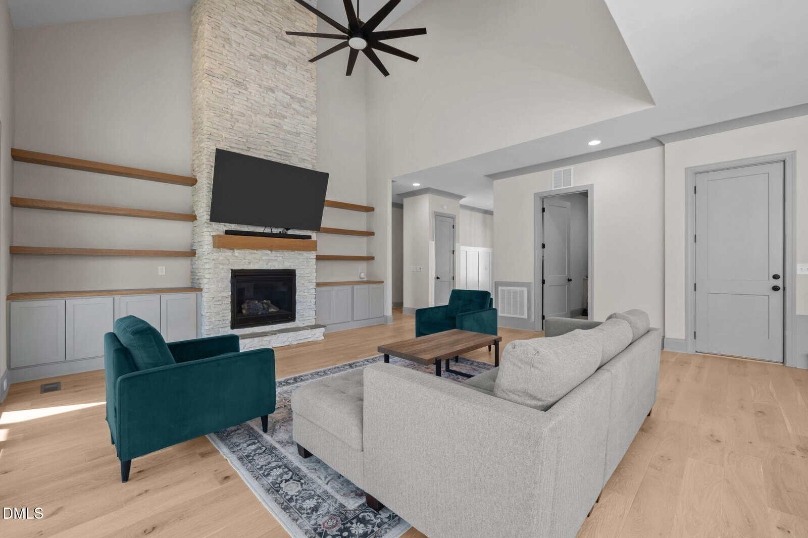 A modern living room with light wood floors, a gray sofa, two teal armchairs, a stone fireplace, a mounted TV, built-in shelves, and a high ceiling with a large black ceiling fan.