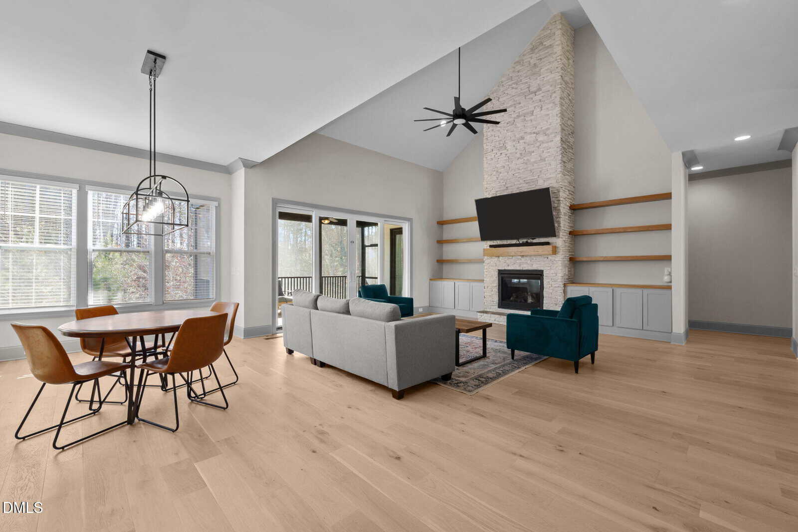 Modern open living and dining area with light wood floors, a stone fireplace with a TV above, built-in shelves, gray sofa, blue armchair, and large windows letting in natural light.