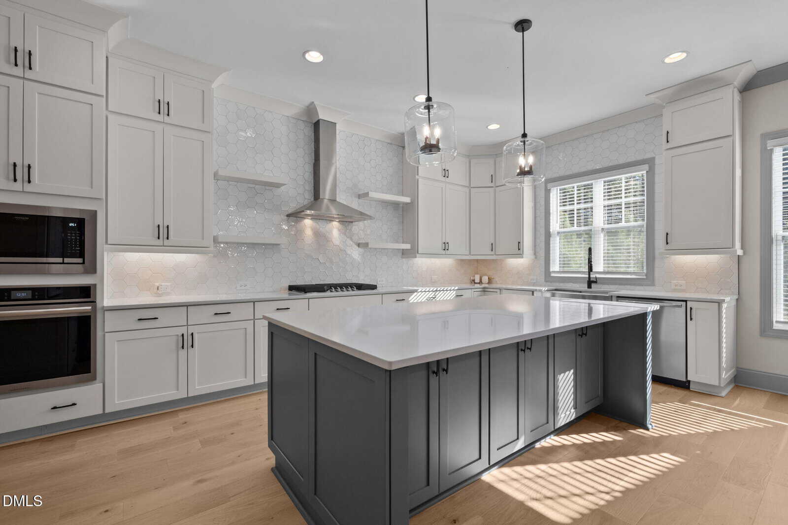 Modern kitchen with white cabinets, hexagonal tile backsplash, stainless steel appliances, a large island with a dark base, pendant lights above, and light wood flooring. Sunlight streams in through two large windows.