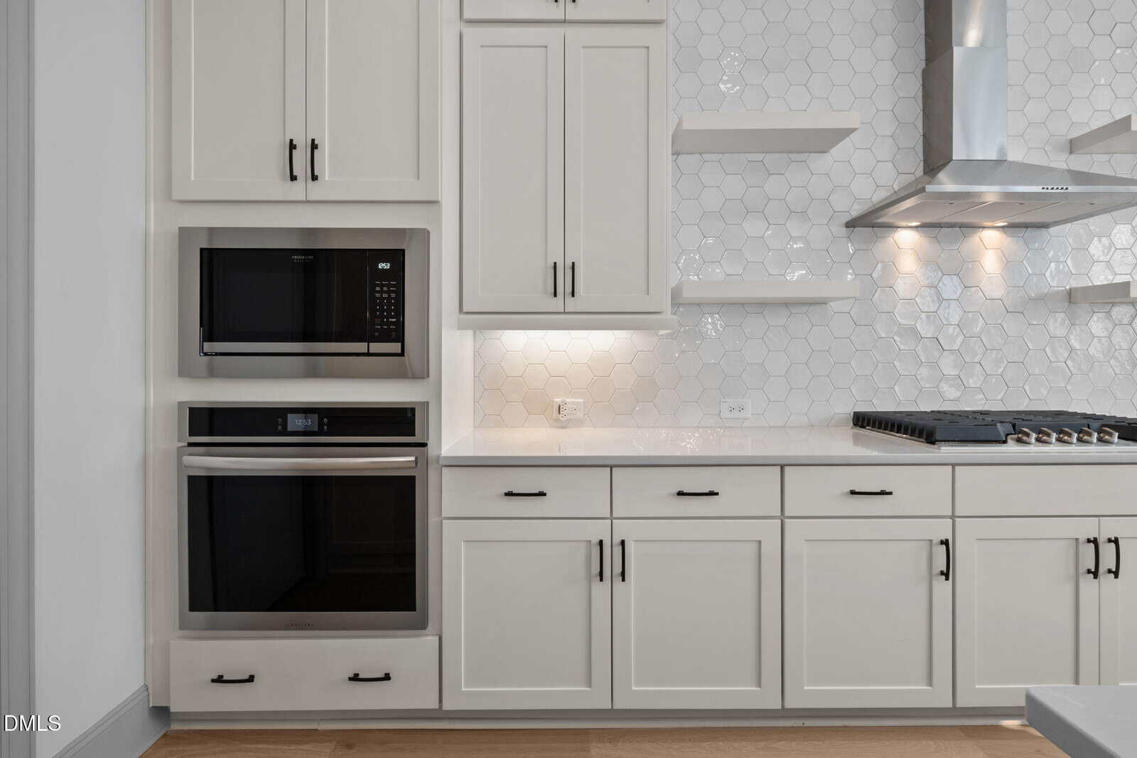 Modern kitchen with white cabinets, black handles, built-in microwave and oven, white hexagon tile backsplash, open shelves, and a stovetop with a stainless steel hood.