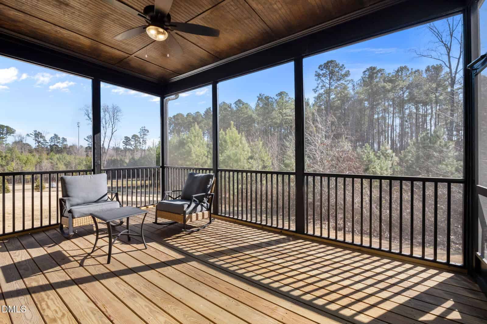 A screened porch with wooden flooring, two gray cushioned chairs, a small table, a ceiling fan, and a view of trees and blue sky in the background. Sunlight casts shadows from the railing onto the floor.