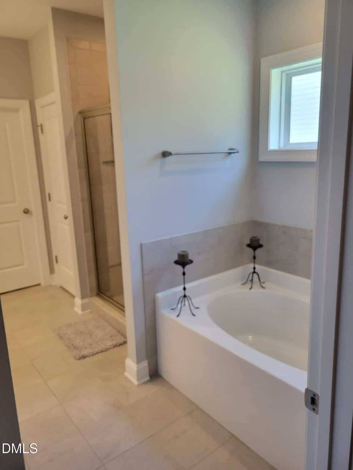 A bright bathroom with a corner bathtub, two candle holders on the tub edge, a towel rack above, a window, and a glass-enclosed shower in the background.