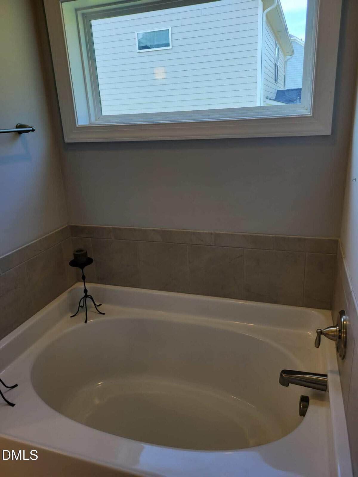 White bathtub with beige tile surround beneath a large window. A black metal candle holder with a candle is placed on the tub’s edge. The window shows the exterior of a nearby house.