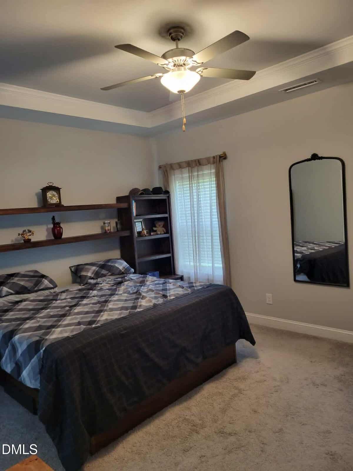 Bedroom with a double bed, gray plaid bedding, wall shelves holding decor, a tall window with sheer curtains, a wall-mounted mirror, and a ceiling fan with lights above. Carpeted floor, neutral walls, and natural light.