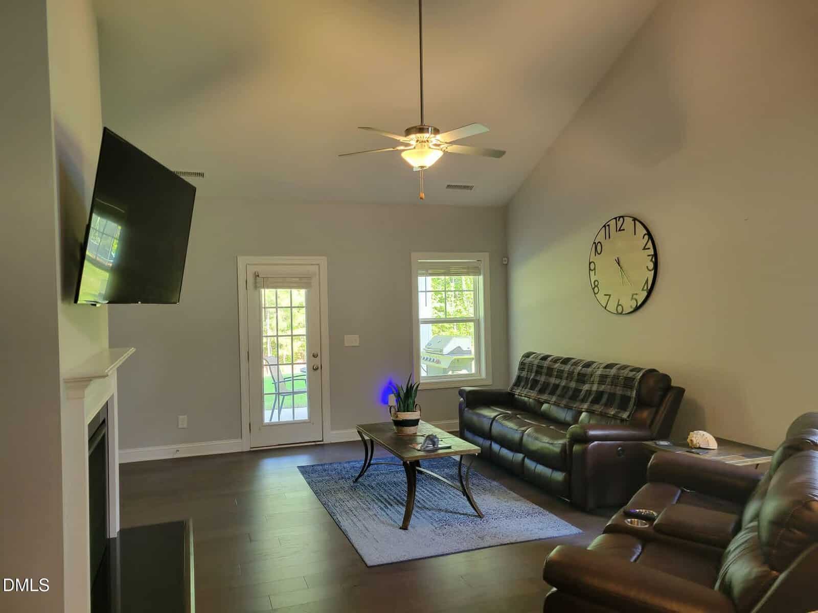 A modern living room with dark leather sofas, a large wall clock, a mounted TV, a ceiling fan, and a coffee table on a rug. Sunlight streams in through two windows and a glass door leading outside.