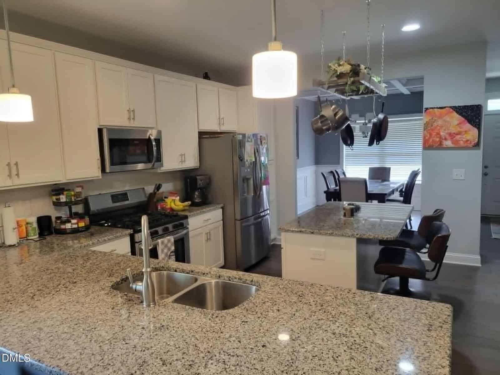 Modern kitchen with granite countertops, white cabinets, stainless steel appliances, a center island with bar stools, hanging pots and pans, and a dining area visible in the background.