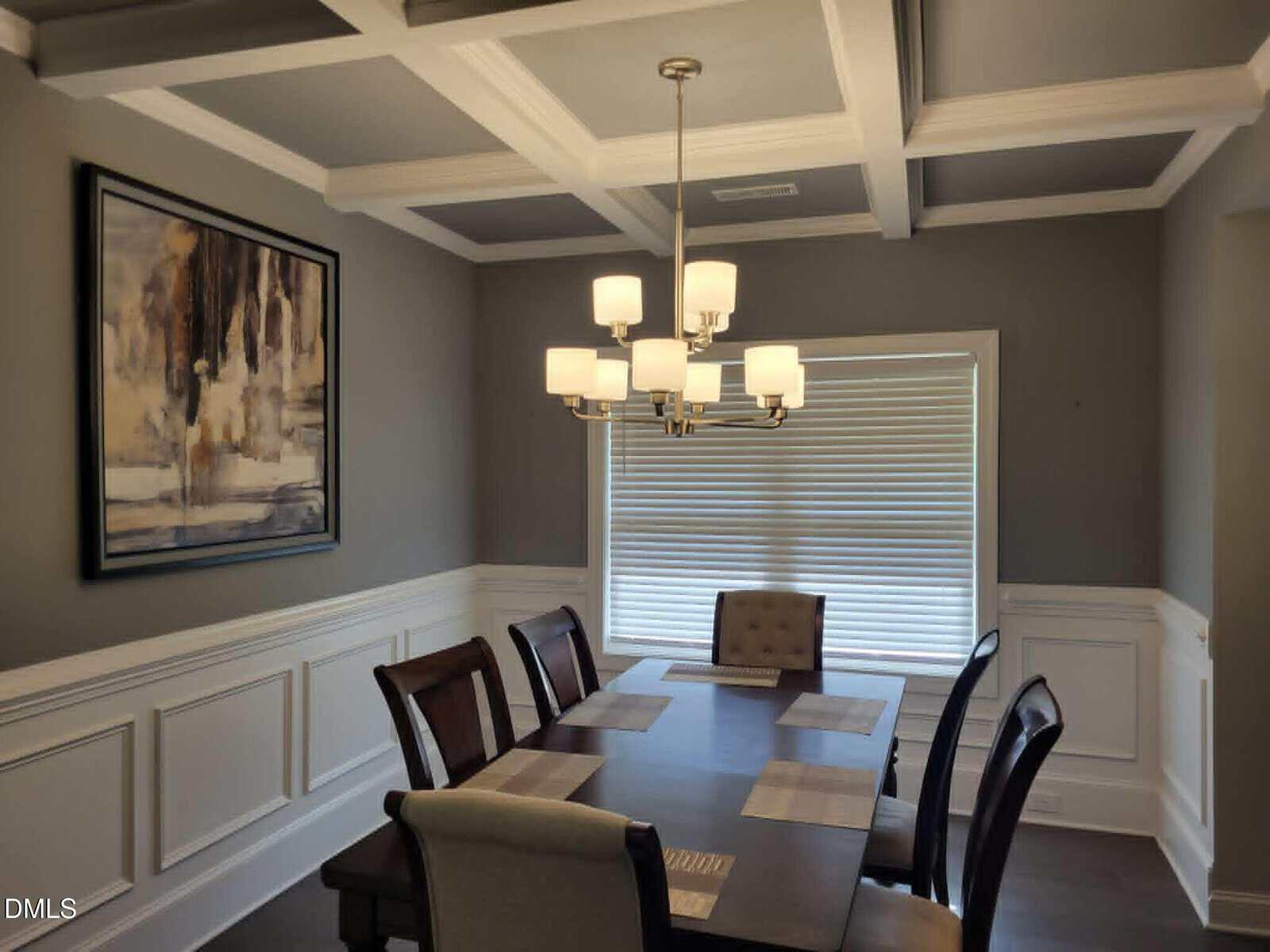 A dining room with a rectangular table and eight chairs, modern chandelier, gray walls with white wainscoting, coffered ceiling, abstract wall art, and a large window with closed blinds.