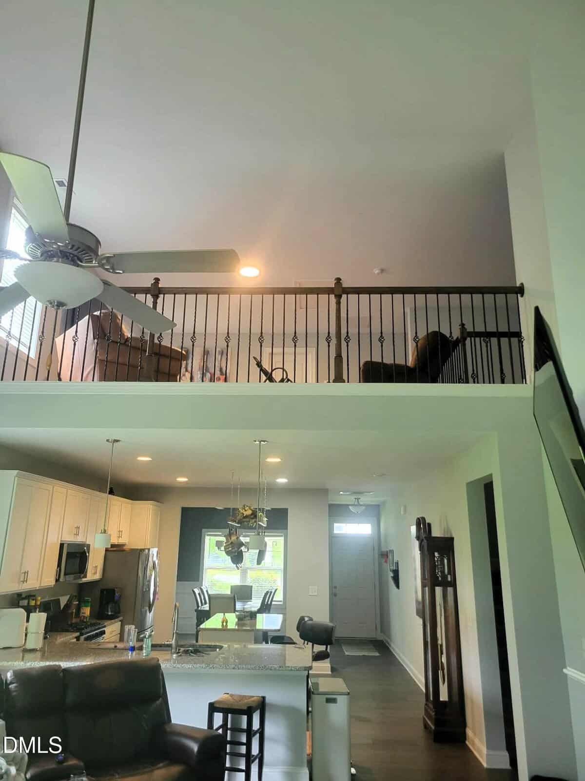 A two-story interior features a kitchen and living area below, with modern appliances and a kitchen island. An open loft area with railing, ceiling fan, and chairs is visible above. A grandfather clock stands near the entryway.