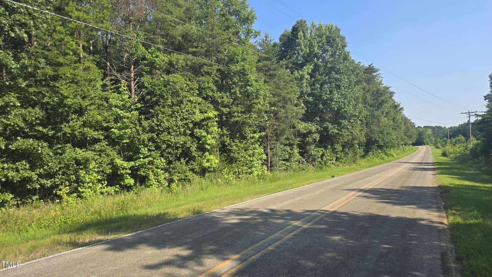 A two-lane rural road bordered by dense green trees and grass under a clear blue sky, with power lines running alongside the road.