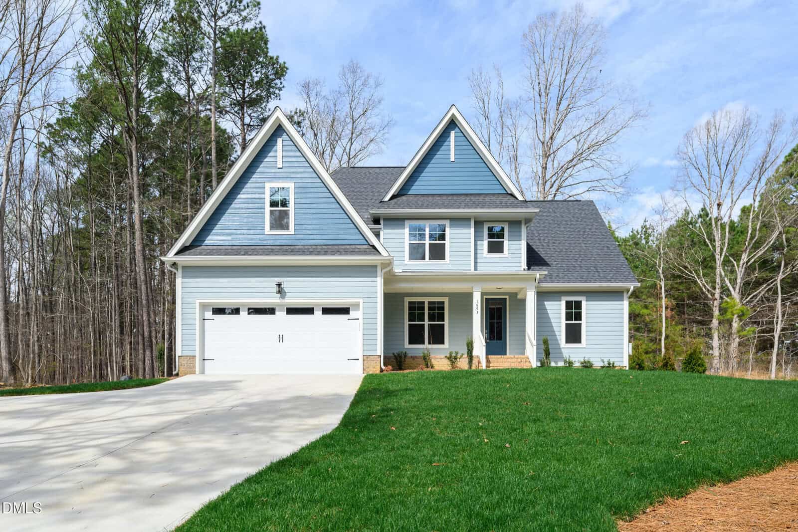 A modern two-story blue house with white trim at 1693 Will Suitt Road in Butner, featuring a double garage and large driveway, surrounded by a grassy lawn and trees under a partly cloudy sky.