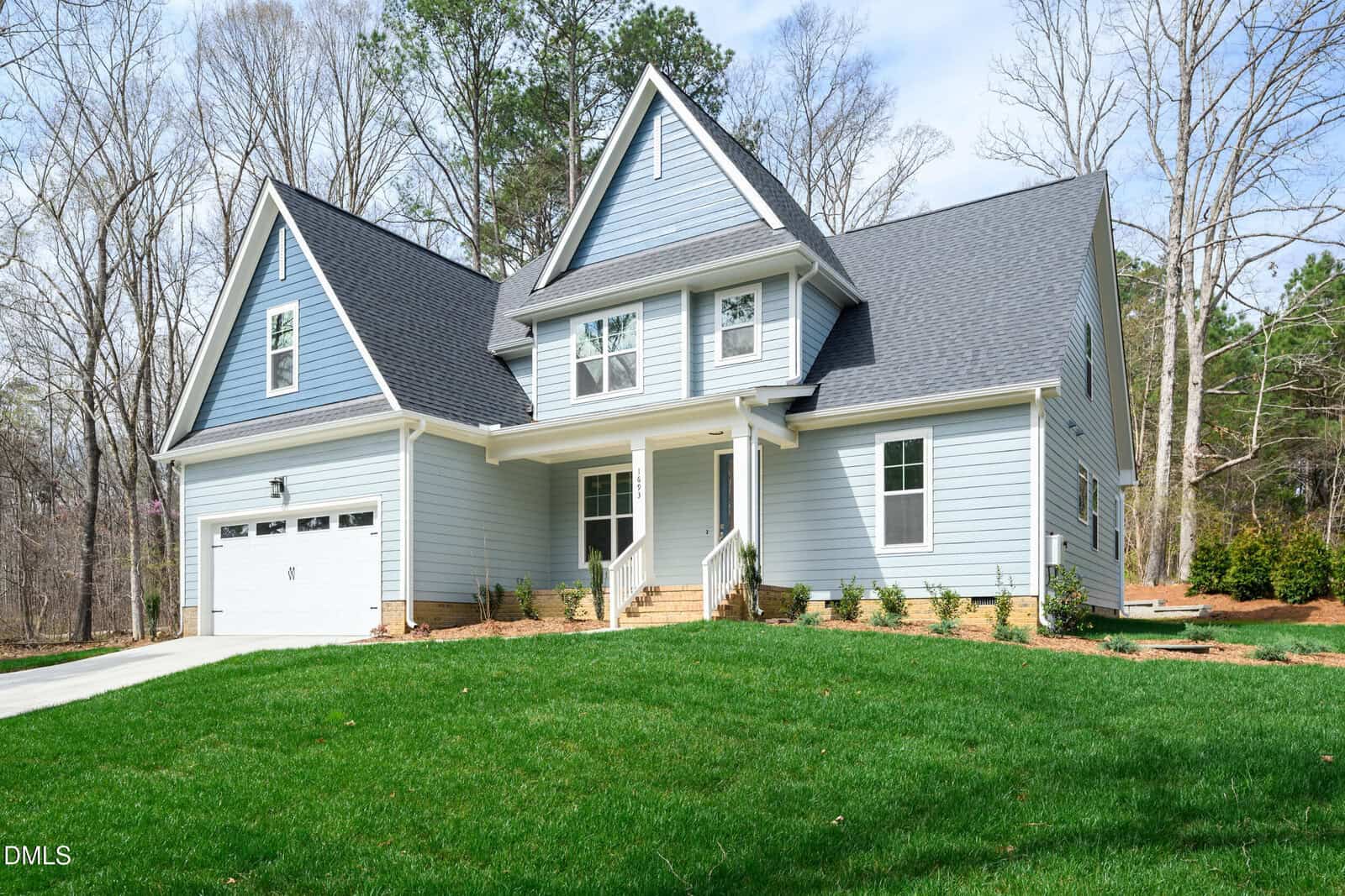 A modern two-story house with light blue siding, white trim, and a dark gray roof. The home has a front porch, a two-car garage, green lawn, and is surrounded by trees on a sunny day.