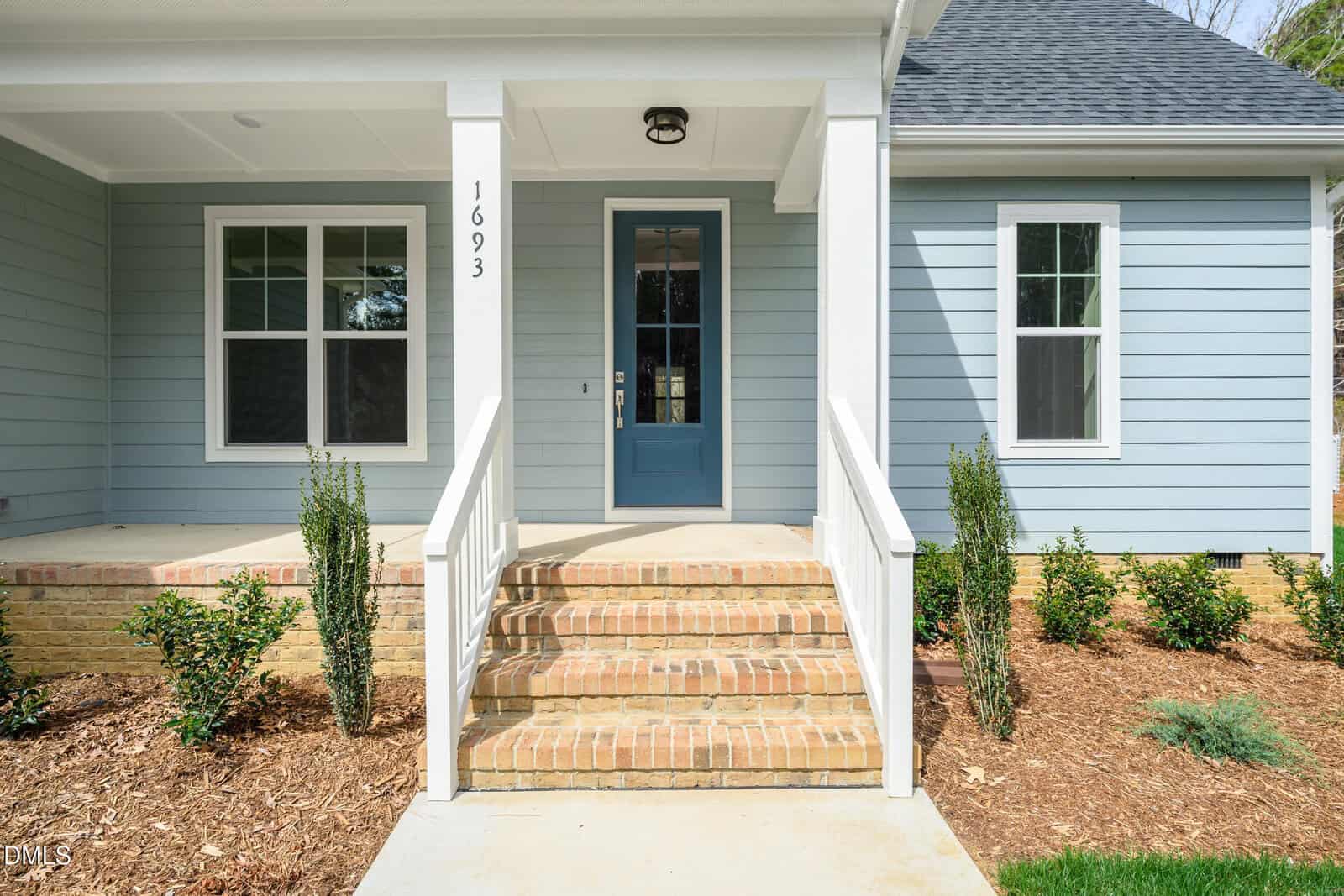 Front entrance of a house with light blue siding, a blue door, white trim, brick steps, white railing, and small shrubs and plants along the mulched walkway.
