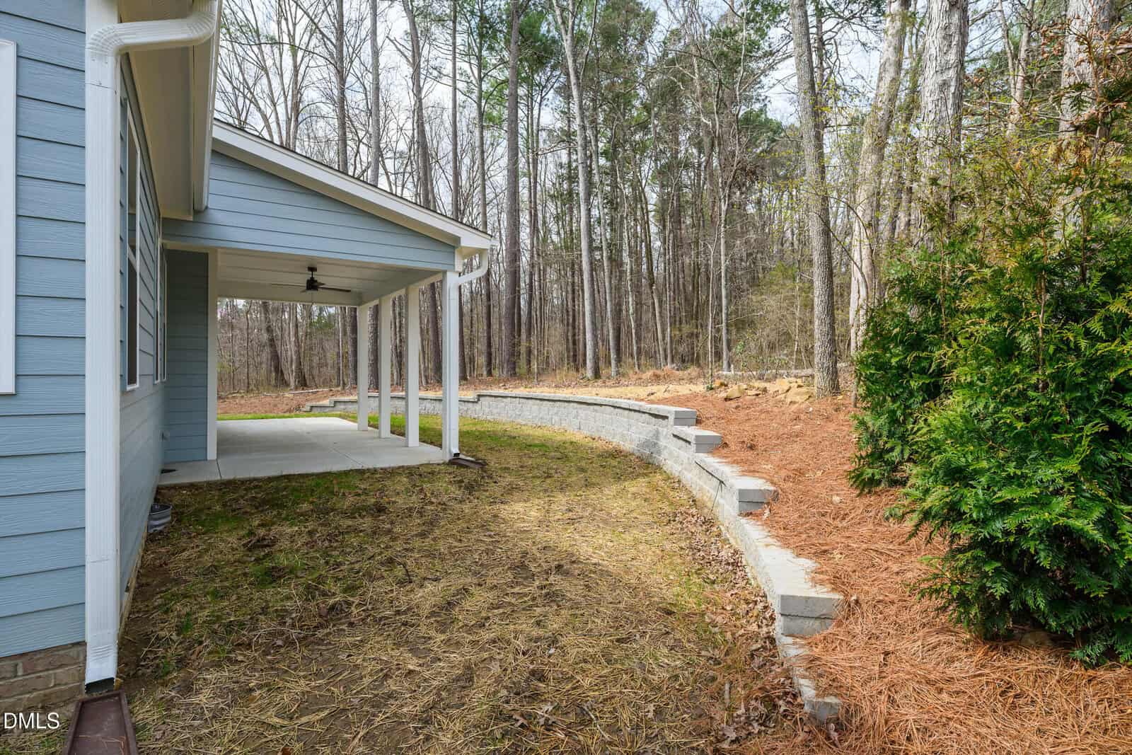 A covered patio with a concrete floor is attached to a blue house, beside a curved stone retaining wall and a wooded backyard with pine straw ground cover.