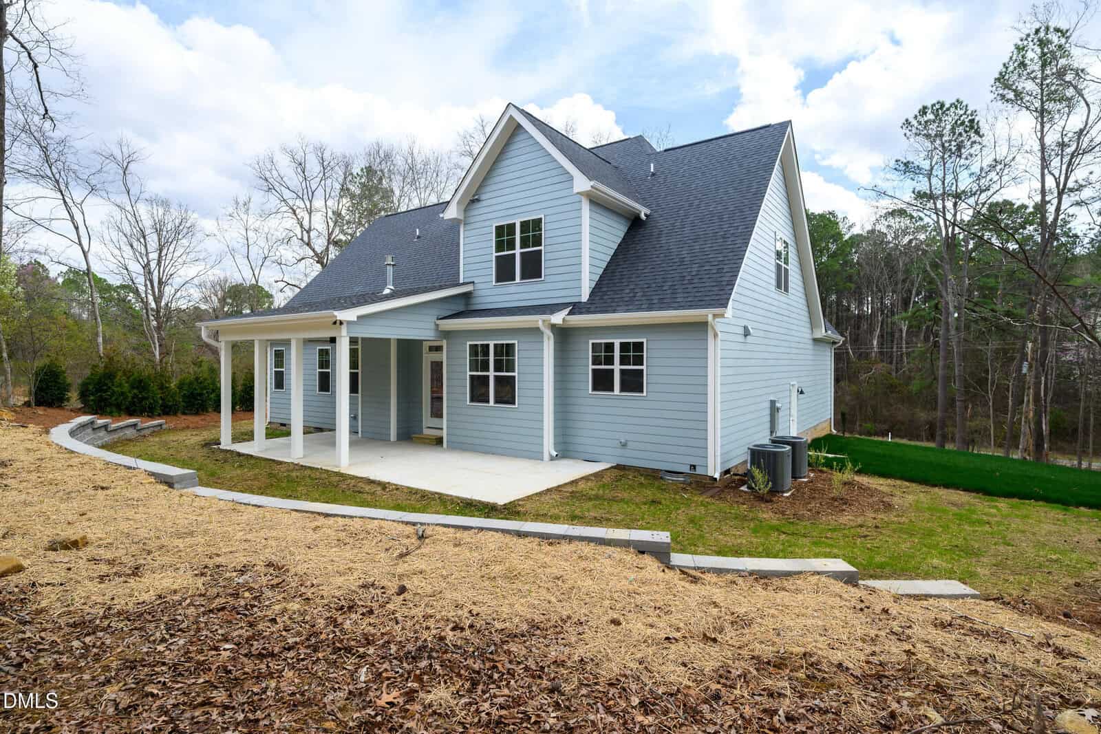 A light blue two-story house with white trim, a covered patio, and multiple windows sits on a sloped yard with mulch and grass, surrounded by trees under a partly cloudy sky.