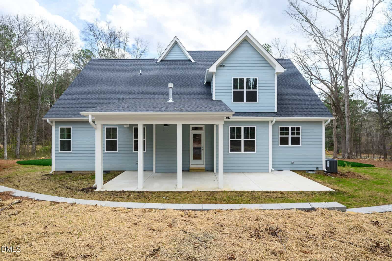 A modern two-story blue house with white trim, a dark roof, and a covered back patio. The yard is partially finished with straw on the ground and surrounded by trees.