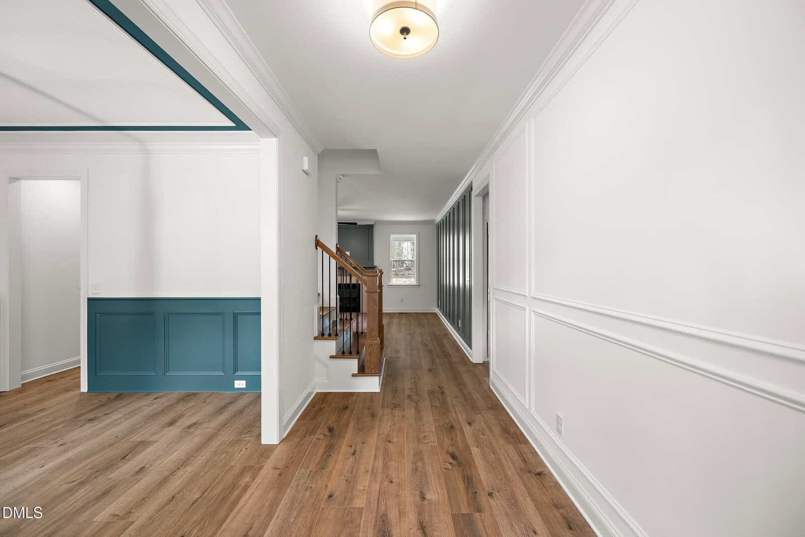 A hallway with wood floors, white walls with trim, a staircase with wooden steps and railing, and a room with teal wainscoting on the left. A ceiling light fixture is visible.