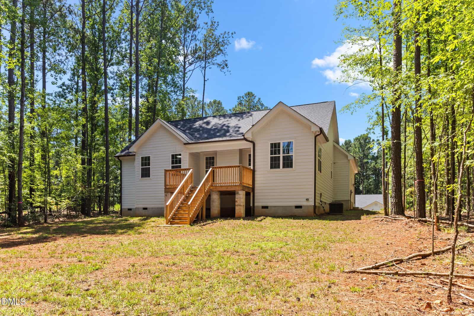 A white single-story house with a small wooden deck and stairs in the backyard, surrounded by tall trees and a grassy, partially shaded yard under a clear blue sky.