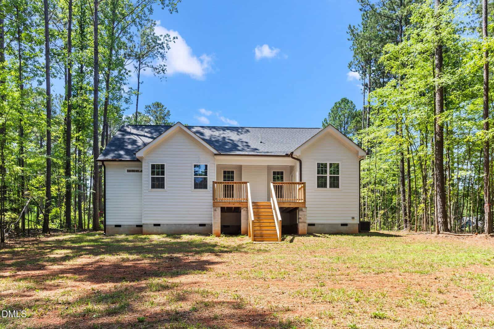 A white, single-story house with a dark roof, wooden steps, and a small porch sits in a grassy clearing surrounded by tall trees under a blue sky with scattered clouds.