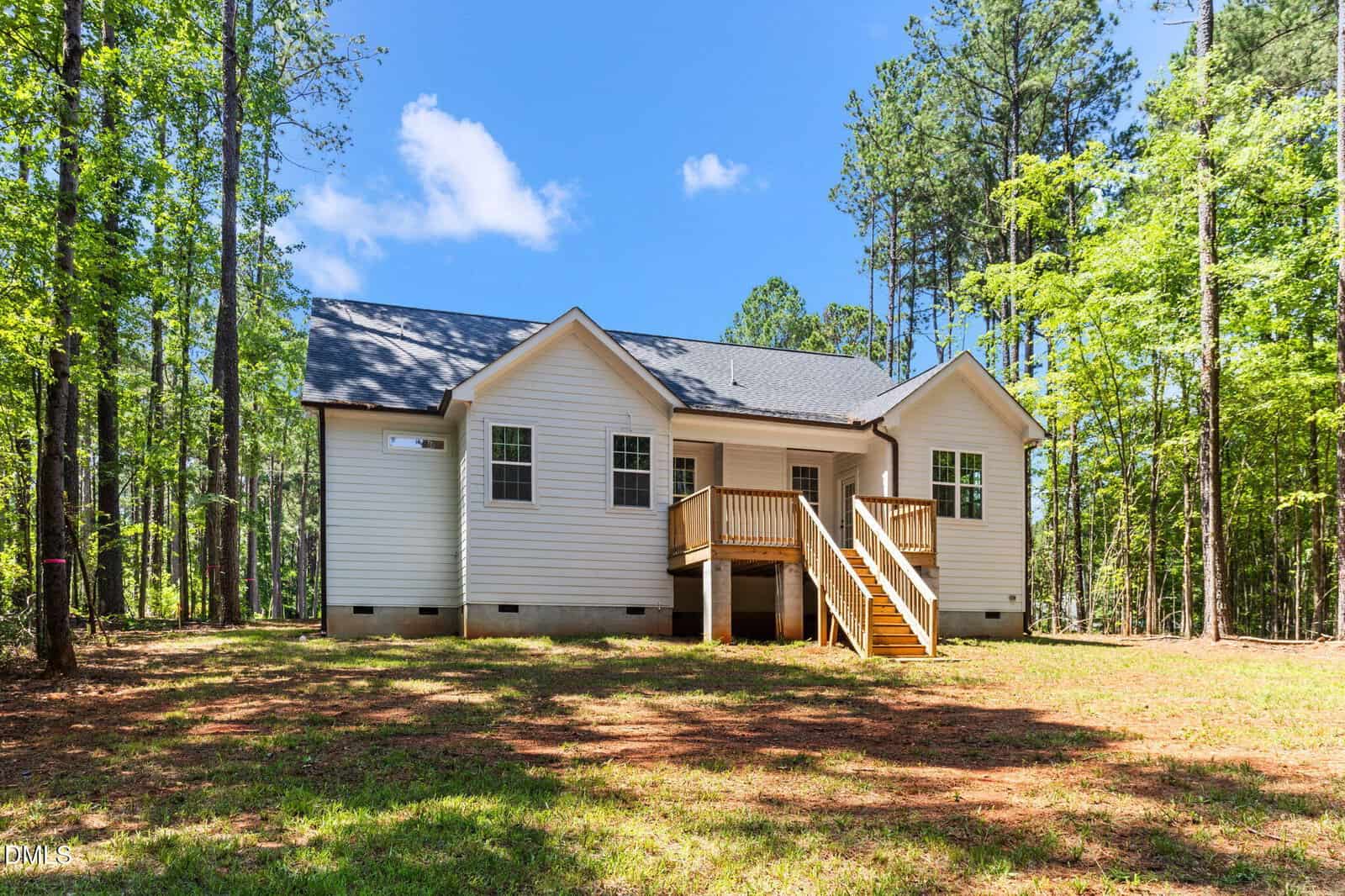 A small, white, single-story house with a dark roof, front porch, and wooden steps, surrounded by tall trees on a sunny day with blue sky and scattered clouds.