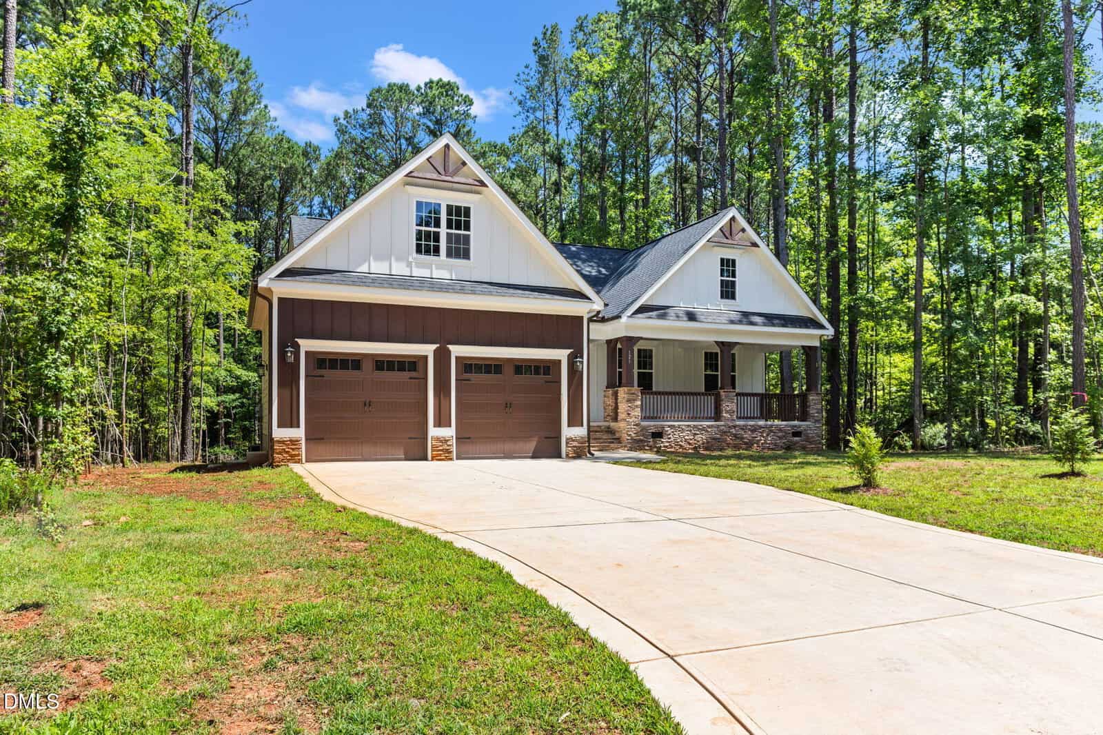 A modern house with two garage doors, a covered front porch, and a large driveway, surrounded by trees and green grass under a blue sky with scattered clouds.