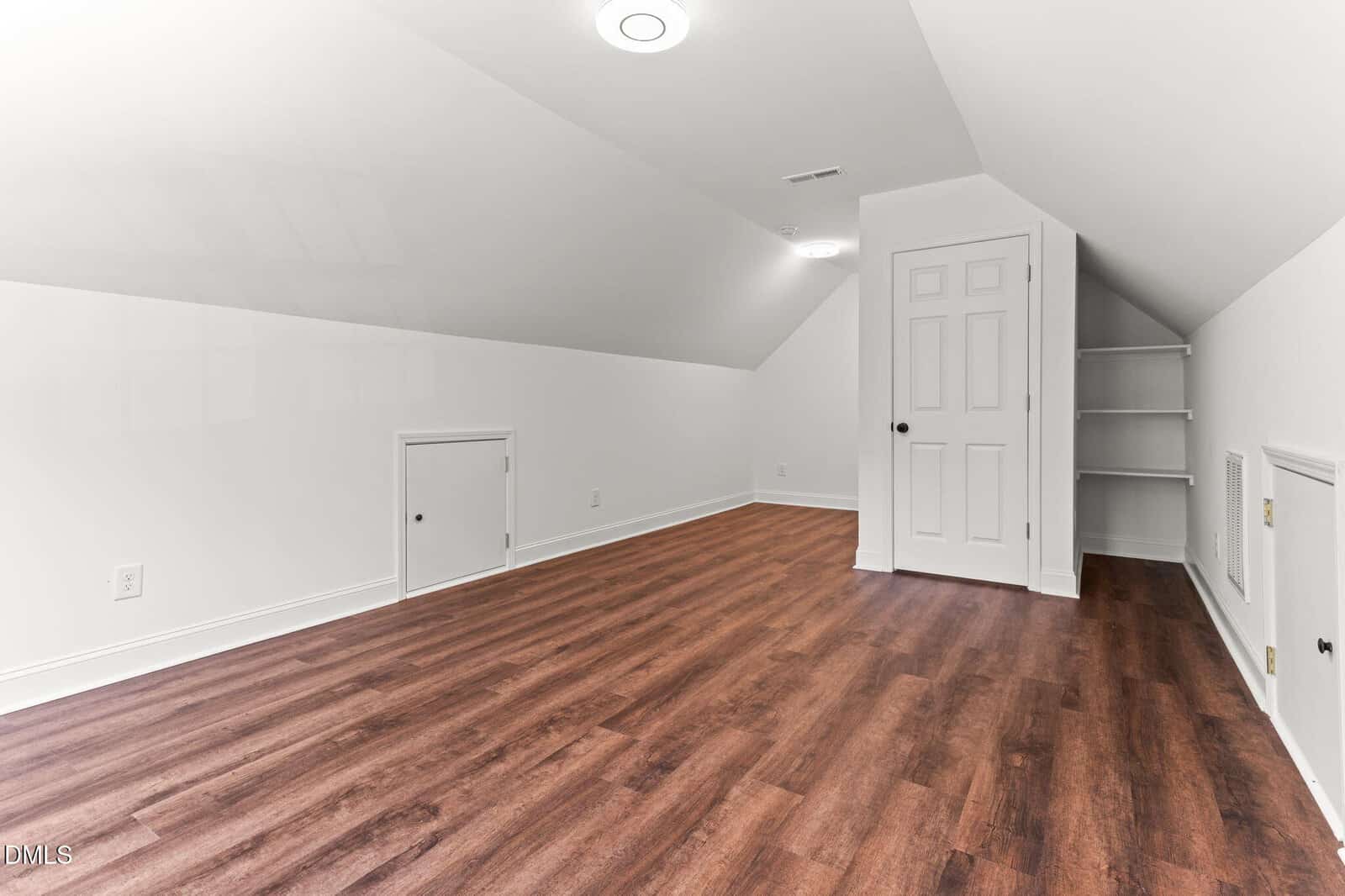 Attic room with sloped white ceiling, recessed lighting, wood-look flooring, a standard door, two small access doors, and built-in corner shelves. No furniture or decorations are present.